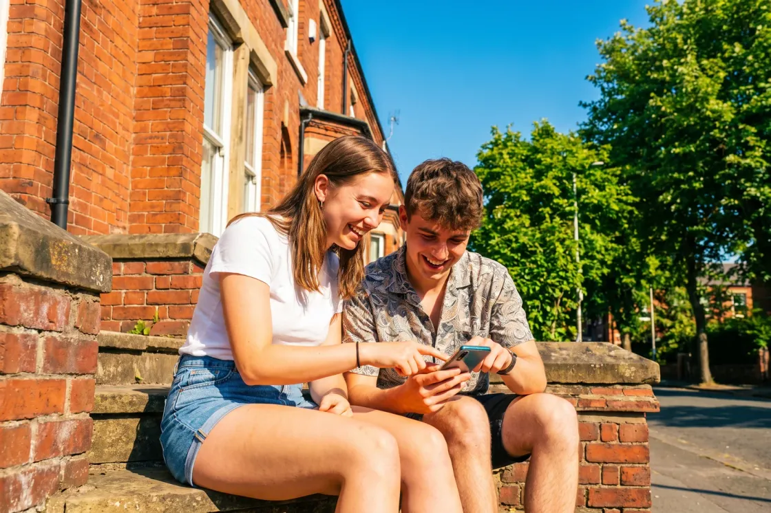 Two students laughing on the sunny front steps of a terraced house while filling in an application on a phone